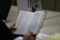 A person is holding an open book, which appears to be a religious text or Bible. The text is in Spanish and is printed in two columns. The person's hands are visible, and there is a Captain America-themed mug and a binder on the table.