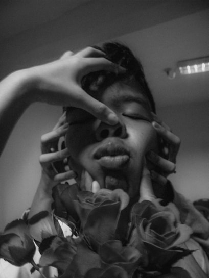 A monochrome image featuring a close-up of a person's face being held by multiple hands, surrounded by roses in the foreground. The lighting casts dramatic shadows, emphasizing the emotional intensity of the composition.