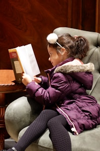 A young girl is sitting in a plush green armchair, engrossed in reading a book. She is wearing a purple coat with a fur-trimmed hood, black patterned tights, and a fluffy headband. Her focus is entirely on the book, which is open in front of her. The setting has a warm wooden interior.