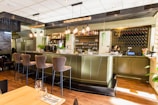 A modern bar featuring a sleek counter with high chairs. The shelves behind the counter are stocked with various bottles of alcohol and glasses. Stylish, hanging pendant lights illuminate the space, with decorative plants adding a touch of greenery. The area is well-organized, and the overall atmosphere is warm and inviting, emphasized by the wooden floor and soft lighting.