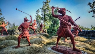 A group of terracotta-colored statues depicting historical warriors in dynamic battle poses. The landscape includes foliage and what appears to be a historical fort wall in the background. The statues are placed on grass-covered mounds, and each figure is armed with traditional weapons, including swords and shields.