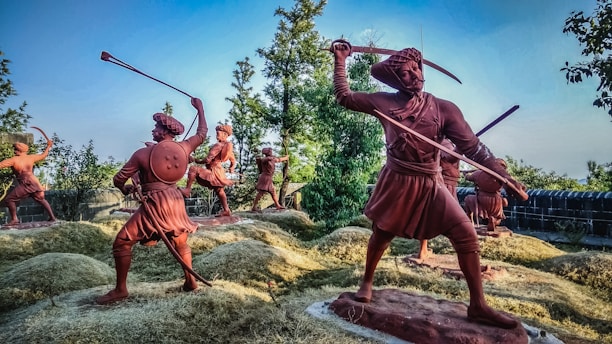 A group of terracotta-colored statues depicting historical warriors in dynamic battle poses. The landscape includes foliage and what appears to be a historical fort wall in the background. The statues are placed on grass-covered mounds, and each figure is armed with traditional weapons, including swords and shields.