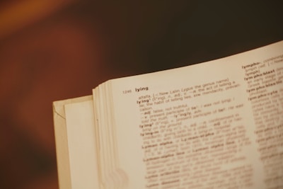 A close-up of an open dictionary page showing the entry for the word 'lying,' including its pronunciation and definitions. The page is slightly yellowed, and the background is blurred, giving a vintage aesthetic.