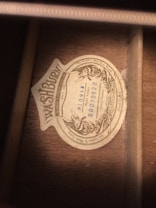 A vintage label is affixed to a wooden surface, bearing the brand name 'Washburn' along with various inscriptions, including 'The World's Best Tone Woods' and serial numbers. The label features ornate, classical design elements around its border.