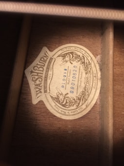 A vintage label is affixed to a wooden surface, bearing the brand name 'Washburn' along with various inscriptions, including 'The World's Best Tone Woods' and serial numbers. The label features ornate, classical design elements around its border.