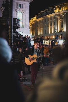 A performer plays the guitar and sings into a microphone on a busy city street at night. The scene is lively with an audience gathered around, including people standing and sitting by ornate architecture. The buildings in the background are illuminated, adding warmth to the night setting.