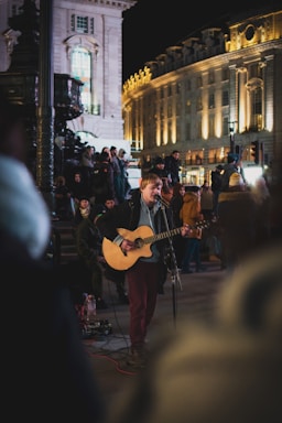 A performer plays the guitar and sings into a microphone on a busy city street at night. The scene is lively with an audience gathered around, including people standing and sitting by ornate architecture. The buildings in the background are illuminated, adding warmth to the night setting.