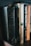 Several books stand side-by-side on a wooden shelf. The titles visible include religious and fiction genres, with one book titled 'The Living Bible' having a notably worn black cover. Other books have spines with dark, beige, and orange tones, and are authored by popular writers.