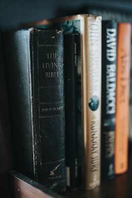 Several books stand side-by-side on a wooden shelf. The titles visible include religious and fiction genres, with one book titled 'The Living Bible' having a notably worn black cover. Other books have spines with dark, beige, and orange tones, and are authored by popular writers.
