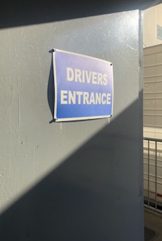 A blue sign with white text reading 'DRIVERS ENTRANCE' attached to a gray wall. Sunlight casts a shadow diagonally across the wall, creating a contrast between light and dark areas. In the background, there is a portion of a white structure with a number partially visible.