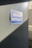 A blue sign with white text reading 'DRIVERS ENTRANCE' attached to a gray wall. Sunlight casts a shadow diagonally across the wall, creating a contrast between light and dark areas. In the background, there is a portion of a white structure with a number partially visible.