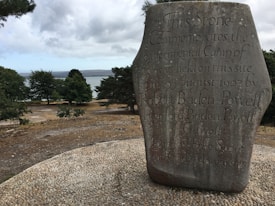 A large stone monument with inscriptions commemorating an experimental camp held in August 1907 by Robert Baden-Powell. The stone is situated in a natural outdoor setting overlooking a body of water surrounded by trees. The sky is overcast with scattered clouds.