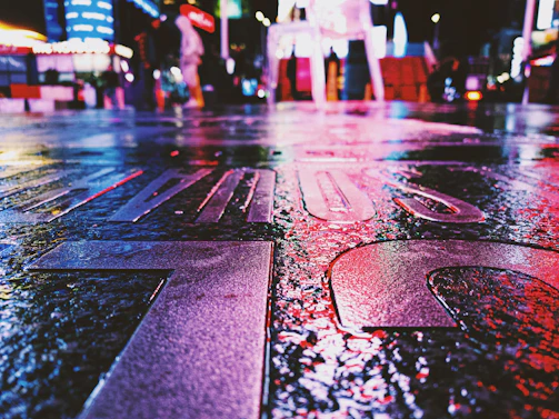 An urban street scene at night, with colorful neon lights reflecting on wet pavement.