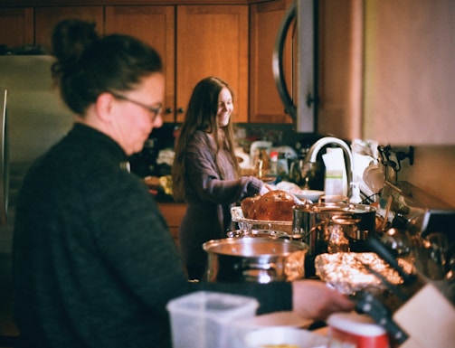 Two people are in a kitchen preparing food. The kitchen is filled with various pots, pans, and a cooked turkey. One person with long hair is smiling and appears to be working at the countertop, while the other is blurred in the foreground, busy with meal preparation.