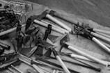 A variety of metal and wooden farming tools are displayed on a surface. There are several hoes and metal implements with wooden handles. Various hooks and rings are shown on the left side. The scene is in black and white, adding a vintage or classic feel.