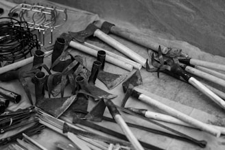 A variety of metal and wooden farming tools are displayed on a surface. There are several hoes and metal implements with wooden handles. Various hooks and rings are shown on the left side. The scene is in black and white, adding a vintage or classic feel.