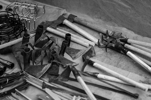 A variety of metal and wooden farming tools are displayed on a surface. There are several hoes and metal implements with wooden handles. Various hooks and rings are shown on the left side. The scene is in black and white, adding a vintage or classic feel.