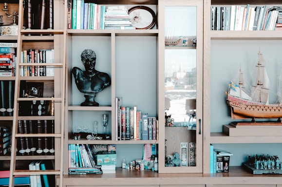 A wooden bookshelf filled with a variety of books, a black bust sculpture, a model ship, and other decorative items. There is a ladder leaning against the shelf. The shelf is neatly organized with books of different colors and sizes, and glass door sections reveal some collectibles inside.