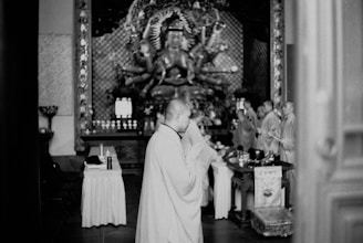 A group of monks is engaged in a religious ceremony inside a temple. The focal point is a monk in the foreground, hands clasped in prayer. In the background, a large multi-armed statue can be seen. The setting appears formal, with elements such as an altar and religious artifacts present.