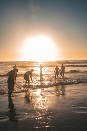 Silhouetted people enjoying the beach as the sun sets on the horizon, casting a warm glow over the ocean and wet sand.