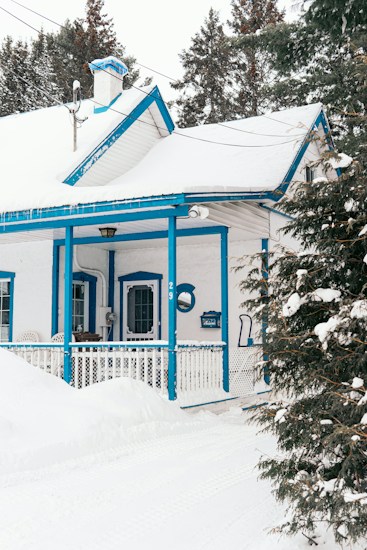 A charming house covered in snow, featuring bright blue accents around the windows, doors, and porch. The surrounding area is blanketed in snow, with a visible snow shovel and some snow-covered trees in the background.