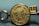 A close-up view of a gold medallion with embossed text and symbols. The text appears around the edge of the medallion and includes words like 'SCHOLASTIC' and 'AWARDS'. The central design features a depiction of a globe and stylized sun rays.