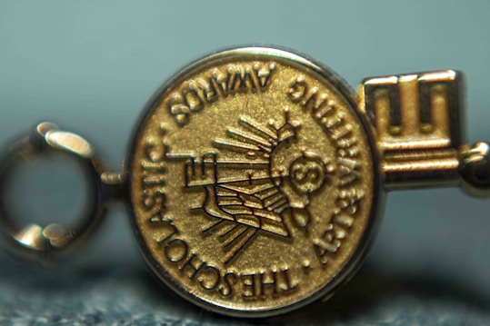 A close-up view of a gold medallion with embossed text and symbols. The text appears around the edge of the medallion and includes words like 'SCHOLASTIC' and 'AWARDS'. The central design features a depiction of a globe and stylized sun rays.
