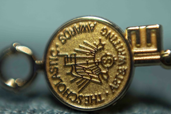 A close-up view of a gold medallion with embossed text and symbols. The text appears around the edge of the medallion and includes words like 'SCHOLASTIC' and 'AWARDS'. The central design features a depiction of a globe and stylized sun rays.