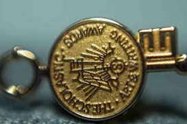 A close-up view of a gold medallion with embossed text and symbols. The text appears around the edge of the medallion and includes words like 'SCHOLASTIC' and 'AWARDS'. The central design features a depiction of a globe and stylized sun rays.