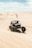 A dune buggy is racing across expansive sand dunes under a partly cloudy sky. A small flag on the vehicle flutters in the breeze, while the sand is kicked up by the spinning wheels.