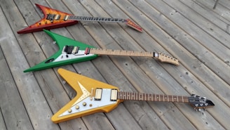 Three electric guitars are laid out on a wooden deck. Each guitar has a distinct color: one red with a flame design, one green with white accents, and one yellow with cream accents. They are arranged in a diagonal line, creating a visually appealing composition.