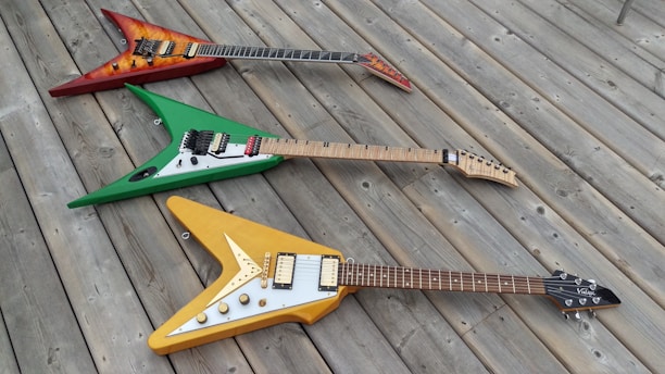 Three electric guitars are laid out on a wooden deck. Each guitar has a distinct color: one red with a flame design, one green with white accents, and one yellow with cream accents. They are arranged in a diagonal line, creating a visually appealing composition.