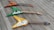 Three electric guitars are laid out on a wooden deck. Each guitar has a distinct color: one red with a flame design, one green with white accents, and one yellow with cream accents. They are arranged in a diagonal line, creating a visually appealing composition.