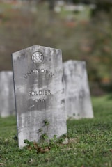 A weathered, gray headstone stands on grass, marked with the text 'UNKNOWN SOLDIER CSA'. In the background, more headstones are visible, creating a serene and somber atmosphere.