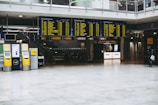 An airport terminal with large electronic flight information display boards showing departure times. Yellow self-service kiosks and signage for Lufthansa are visible, with a sparse number of people in the background.