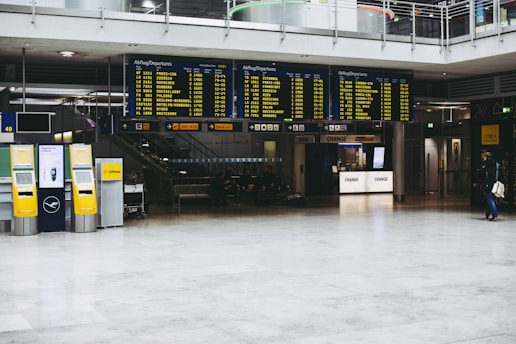 An airport terminal with large electronic flight information display boards showing departure times. Yellow self-service kiosks and signage for Lufthansa are visible, with a sparse number of people in the background.