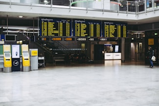 An airport terminal with large electronic flight information display boards showing departure times. Yellow self-service kiosks and signage for Lufthansa are visible, with a sparse number of people in the background.