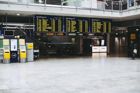 An airport terminal with large electronic flight information display boards showing departure times. Yellow self-service kiosks and signage for Lufthansa are visible, with a sparse number of people in the background.
