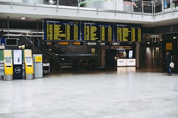 An airport terminal with large electronic flight information display boards showing departure times. Yellow self-service kiosks and signage for Lufthansa are visible, with a sparse number of people in the background.