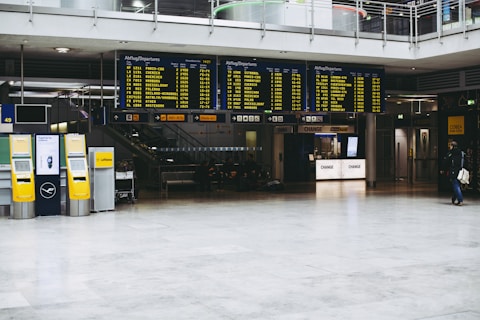 An airport terminal with large electronic flight information display boards showing departure times. Yellow self-service kiosks and signage for Lufthansa are visible, with a sparse number of people in the background.