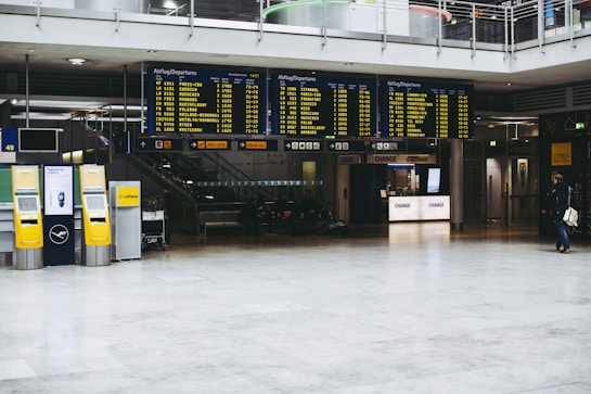 An airport terminal with large electronic flight information display boards showing departure times. Yellow self-service kiosks and signage for Lufthansa are visible, with a sparse number of people in the background.
