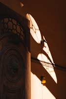 A wooden door with intricate circular patterns is partially illuminated by sunlight, casting shadow patterns on a warm-colored wall. A small security camera and a yellow light fixture are mounted on the wall.