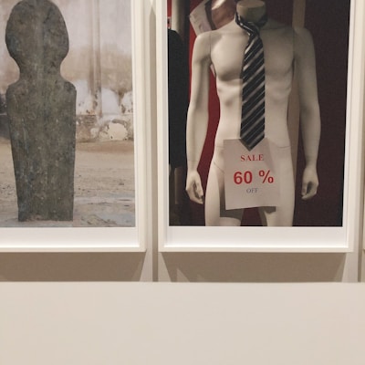 A mannequin is displayed wearing a striped tie and holding a sale sign that reads '60% off'. The scene is minimalistic with a focus on commercialism. Next to it, a textured stone or concrete sculpture with a smooth surface and abstract shape is presented.