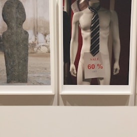 A mannequin is displayed wearing a striped tie and holding a sale sign that reads '60% off'. The scene is minimalistic with a focus on commercialism. Next to it, a textured stone or concrete sculpture with a smooth surface and abstract shape is presented.