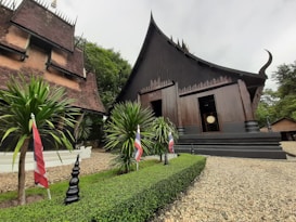 A traditional Thai-style building with a steep, sloping roof is surrounded by lush greenery and gravel pathways. Small trees and plants are neatly arranged alongside the building, with flags placed intermittently along the garden edge. The architecture features intricate woodwork and dark wooden surfaces, complementing the natural surroundings.
