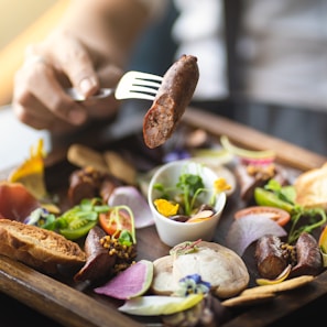 A hand holds a fork with a piece of sausage over a wooden platter filled with an assortment of gourmet foods. The platter includes various meats, fresh greens, sliced vegetables, edible flowers, bread, and a small dipping bowl.