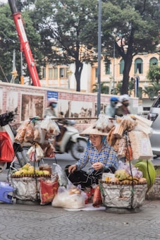 A street vendor wearing a traditional conical hat is surrounded by baskets filled with various goods, including fruits and pre-packaged snacks. The vendor is seated on a sidewalk, with a busy street and blurred motorcycles passing by in the background. Trees and a building facade are also visible.