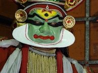 A performer wearing traditional makeup and attire associated with classical Indian dance, characterized by vibrant colors and elaborate headgear. The face is painted in green with bold black and red designs, and the costume includes intricate jewelry and fabric decorations.
