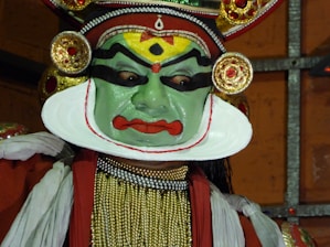 A performer wearing traditional makeup and attire associated with classical Indian dance, characterized by vibrant colors and elaborate headgear. The face is painted in green with bold black and red designs, and the costume includes intricate jewelry and fabric decorations.