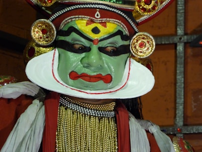 A performer wearing traditional makeup and attire associated with classical Indian dance, characterized by vibrant colors and elaborate headgear. The face is painted in green with bold black and red designs, and the costume includes intricate jewelry and fabric decorations.